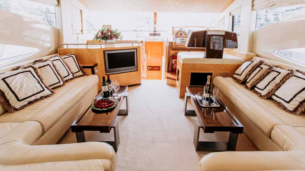 yacht saloon having leather sofa and cushions, saloon windows, wooden tables, built in tv and steering station